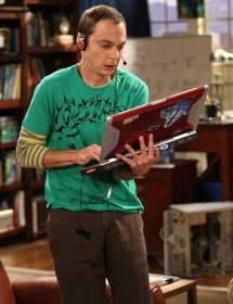 Sheldon de pie, jugando a un videojuego con su portatil con unos auriculares y un micrfono