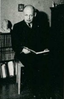 Rudolf Spielmann posa con un libro en sus manos, detrás de él hay una pequeña librería repleta de libros