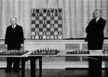 Spielmann de pie frente a un tablero mural que contiene las dos primeras jugadas de un gambito de rey: 1.e4 e5 2.f4 d5. Delante de él tiene 3 juego de ajedrez y a su derecha aparece el organizador del evento.