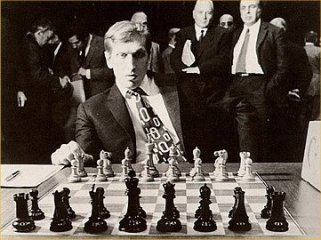 Fischer espera el comienzo de la partida ante el tablero, observando fiajmente las piezas. A su espalda, varias personas le observan